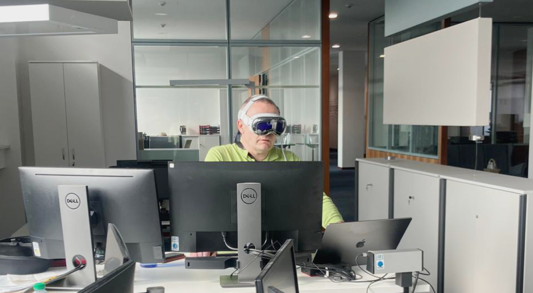 Apple-Vision-Test Eine Mitarbeiter an einem Schreibtisch mit Computermonitoren trägt eine Apple Vision Pro.